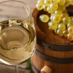 Copa de vino blanco junto a un barril de madera con uvas. El vino, con su crianza sobre lías, promete un sabor único.