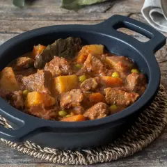 Estofado de ternera con patatas, guisantes y laurel, servido en cazuela negra sobre mesa de madera.