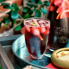 Un refrescante tinto de verano con hielo y rodajas de limón en un vaso, junto a una jarra llena de la misma bebida y un cuenco con palitos de queso.