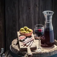 Copa de vino tinto, aceitunas, higo y salchichones sobre una tabla de madera, junto a una jarra de vino. Un festín que evoca la pregunta: ¿qué es el moscatel?