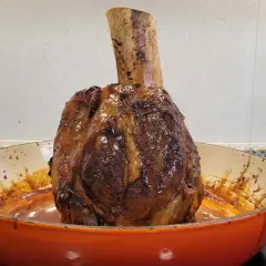Un jugoso jarrete de ternera asado, con su hueso expuesto, reposa en una cazuela naranja, listo para ser disfrutado.