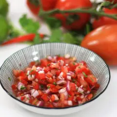 Un tazón de pico de gallo fresco, con tomates, cebolla morada y cilantro picados. Al fondo, tomates, limas y chiles rojos.