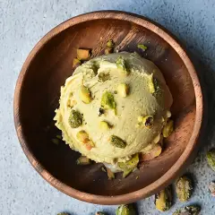 Cremoso helado de pistacho servido en un cuenco de madera, adornado con trozos de pistacho y acompañado de más pistachos enteros esparcidos.