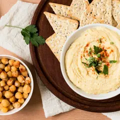 Un tazón de hummus cremoso, decorado con perejil y pimentón, junto a garbanzos y galletas saladas. ¡Perfecto para tu hummus receta!