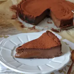 Una porción de tarta de chocolate, cubierta de cacao en polvo, reposa en un plato blanco. El resto de la tarta, también espolvoreada, se ve al fondo.