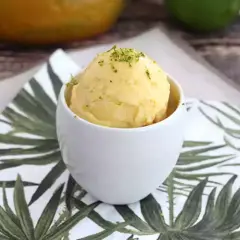 Una bola de sorbete de mango, refrescante y deliciosa, servida en una taza blanca sobre una servilleta con hojas tropicales.