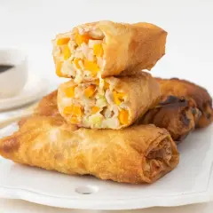 Rollitos de primavera dorados y crujientes, rellenos de verduras y carne. Se apilan sobre una bandeja blanca, con salsa de soja al fondo.