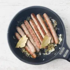 Salchichas al vino dorándose en sartén con cebolla y hojas de laurel.