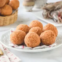 Croquetas de gambas doradas y crujientes en un plato blanco, con más en una cesta de mimbre al fondo.