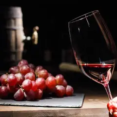 Copa de vino tinto con uvas rojas y un barril al fondo, evocando los taninos del vino.