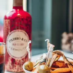Un vaso de vermut con hielo y naranja, junto a una botella roja de Padró & Co. Vermouth y un plato de patatas y anchoas. ¡Perfecto para clases de vermut!