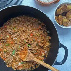 Un delicioso plato de ropa vieja, con trozos de carne deshebrada en salsa roja, acompañado de plátanos maduros fritos y arroz blanco.