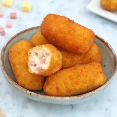 Un tazón lleno de crujientes croquetas de jamón, una de ellas partida mostrando su cremoso interior con trocitos de jamón.