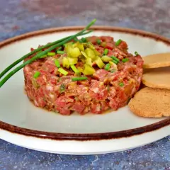 Tartar de salchichón picado finamente, adornado con cebollino y pepinillos, servido con tostas.