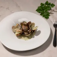 Un plato blanco con almejas en salsa verde, adornado con perejil fresco. Un tenedor y una servilleta completan la escena.