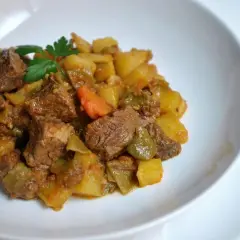 Un plato de carne guisada con patatas, zanahorias y pimientos verdes, adornado con perejil fresco.