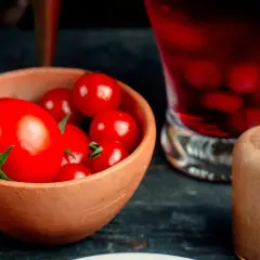 Un cuenco de barro lleno de tomates rojos brillantes, listos para un delicioso tartar de tomate. A su lado, un vaso con bebida roja y un salero de madera.