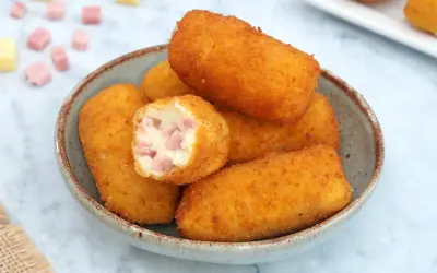 Un tazón lleno de crujientes croquetas de jamón, una de ellas partida mostrando su cremoso interior con trocitos de jamón.