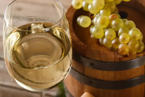 Copa de vino blanco junto a un barril de madera con uvas. El vino, con su crianza sobre lías, promete un sabor único.