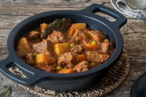 Estofado de ternera con patatas, guisantes y laurel, servido en cazuela negra sobre mesa de madera.