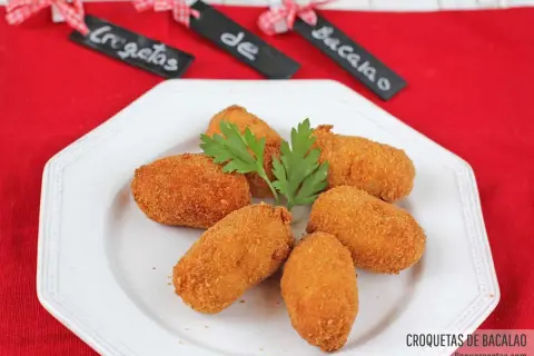 Seis croquetas de bacalao doradas y crujientes, adornadas con perejil fresco, servidas en un plato blanco.