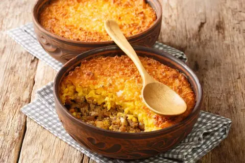 Dos cazuelas de barro con pastel de choclo, una con cuchara de madera. Un plato chileno casero y delicioso.