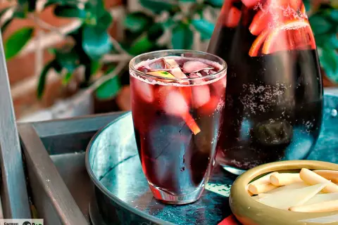 Un refrescante tinto de verano con hielo y rodajas de limón en un vaso, junto a una jarra llena de la misma bebida y un cuenco con palitos de queso.