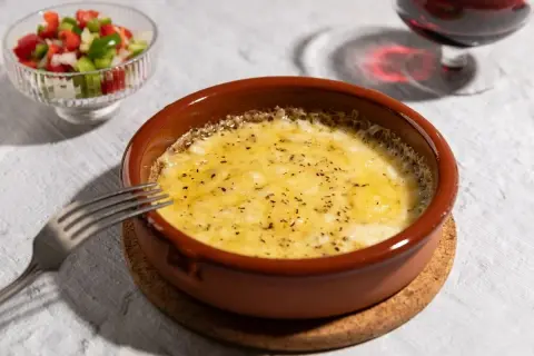 Delicioso provolone al horno burbujeante, servido en cazuela de barro con una guarnición de pimientos y una copa de vino tinto.