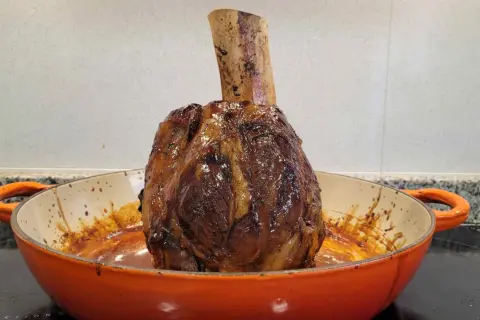 Un jugoso jarrete de ternera asado, con su hueso expuesto, reposa en una cazuela naranja, listo para ser disfrutado.