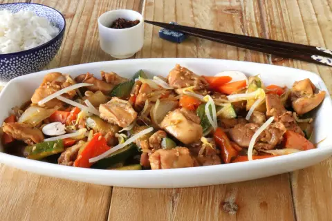Un delicioso chop suey de pollo con verduras y brotes de soja, servido en un plato blanco junto a un tazón de arroz blanco y palillos.