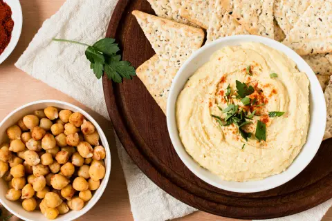 Un tazón de hummus cremoso, decorado con perejil y pimentón, junto a garbanzos y galletas saladas. ¡Perfecto para tu hummus receta!