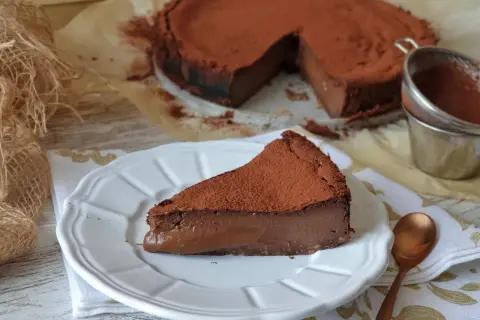 Una porción de tarta de chocolate, cubierta de cacao en polvo, reposa en un plato blanco. El resto de la tarta, también espolvoreada, se ve al fondo.