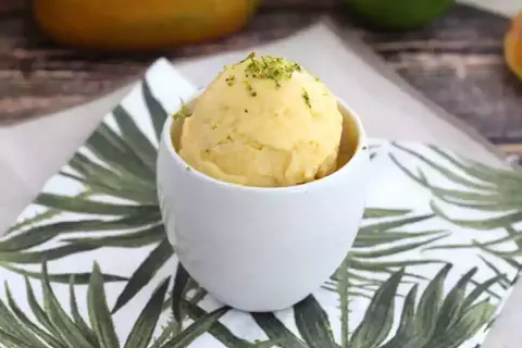 Una bola de sorbete de mango, refrescante y deliciosa, servida en una taza blanca sobre una servilleta con hojas tropicales.