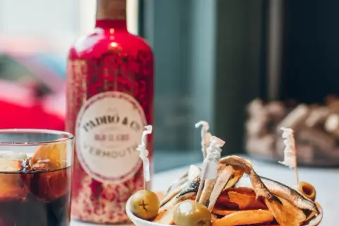 Un vaso de vermut con hielo y naranja, junto a una botella roja de Padró & Co. Vermouth y un plato de patatas y anchoas. ¡Perfecto para clases de vermut!