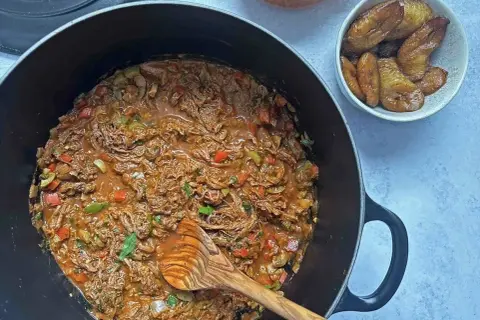 Un delicioso plato de ropa vieja, con trozos de carne deshebrada en salsa roja, acompañado de plátanos maduros fritos y arroz blanco.