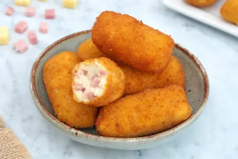 Un tazón lleno de crujientes croquetas de jamón, una de ellas partida mostrando su cremoso interior con trocitos de jamón.