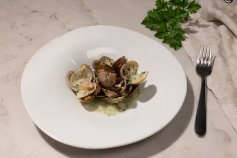 Un plato blanco con almejas en salsa verde, adornado con perejil fresco. Un tenedor y una servilleta completan la escena.