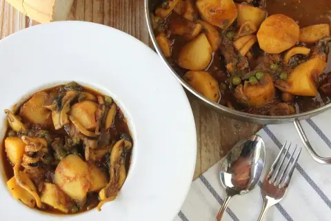 Plato de papas con choco y guisantes en salsa, servido en un cuenco blanco. Al lado, una sartén con el mismo guiso.