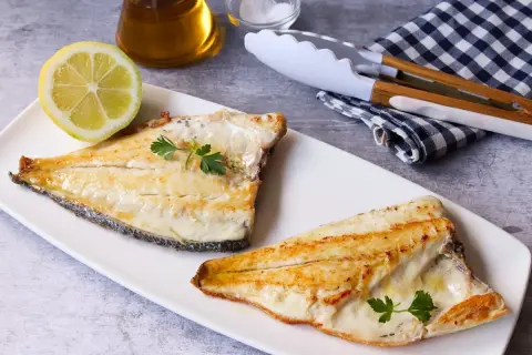 Dos filetes de dorada a la plancha, servidos en plato blanco con limón, perejil y utensilios de cocina.