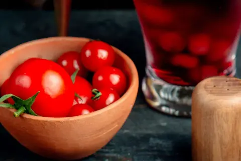Un cuenco de barro lleno de tomates rojos brillantes, listos para un delicioso tartar de tomate. A su lado, un vaso con bebida roja y un salero de madera.