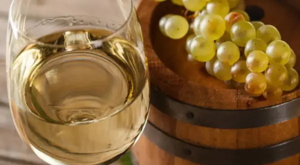 Copa de vino blanco junto a un barril de madera con uvas. El vino, con su crianza sobre lías, promete un sabor único.