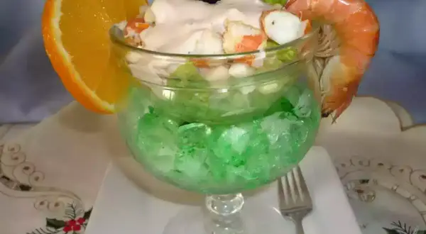 Delicioso coctel de marisco servido en copa con hielo verde, adornado con naranja y gamba.