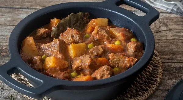 Estofado de ternera con patatas, guisantes y laurel, servido en cazuela negra sobre mesa de madera.