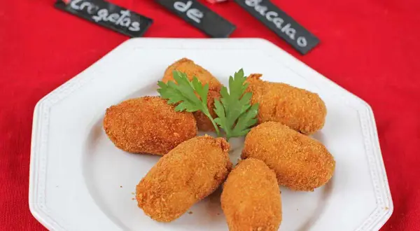 Seis croquetas de bacalao doradas y crujientes, adornadas con perejil fresco, servidas en un plato blanco.