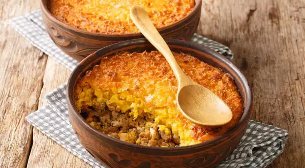 Dos cazuelas de barro con pastel de choclo, una con cuchara de madera. Un plato chileno casero y delicioso.