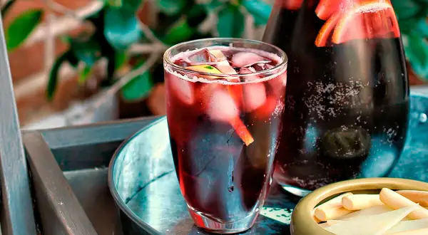 Un refrescante tinto de verano con hielo y rodajas de limón en un vaso, junto a una jarra llena de la misma bebida y un cuenco con palitos de queso.