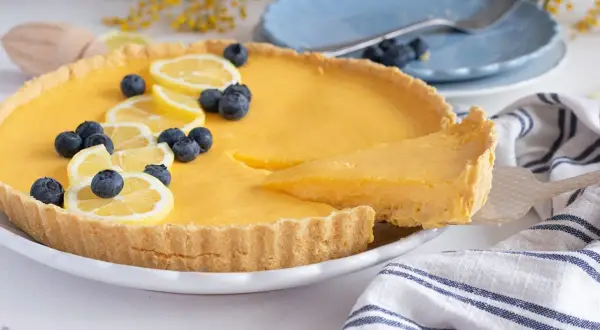 Deliciosa tarta de limón, decorada con rodajas de limón y arándanos. Una porción se sirve con una espátula.