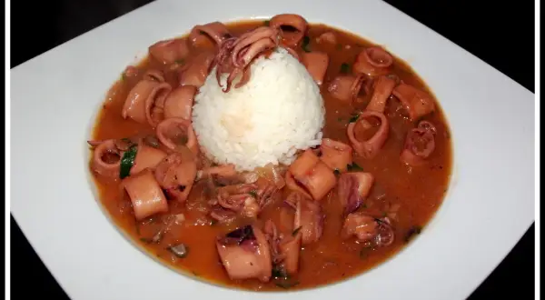 Calamares encebollados en una salsa rojiza, servidos con una bola de arroz blanco en un plato blanco.