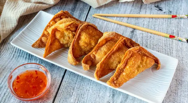 Crujientes wan tun frito dorados en una bandeja blanca, acompañados de salsa agridulce y palillos.