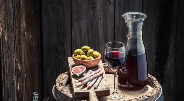 Copa de vino tinto, aceitunas, higo y salchichones sobre una tabla de madera, junto a una jarra de vino. Un festín que evoca la pregunta: ¿qué es el moscatel?
